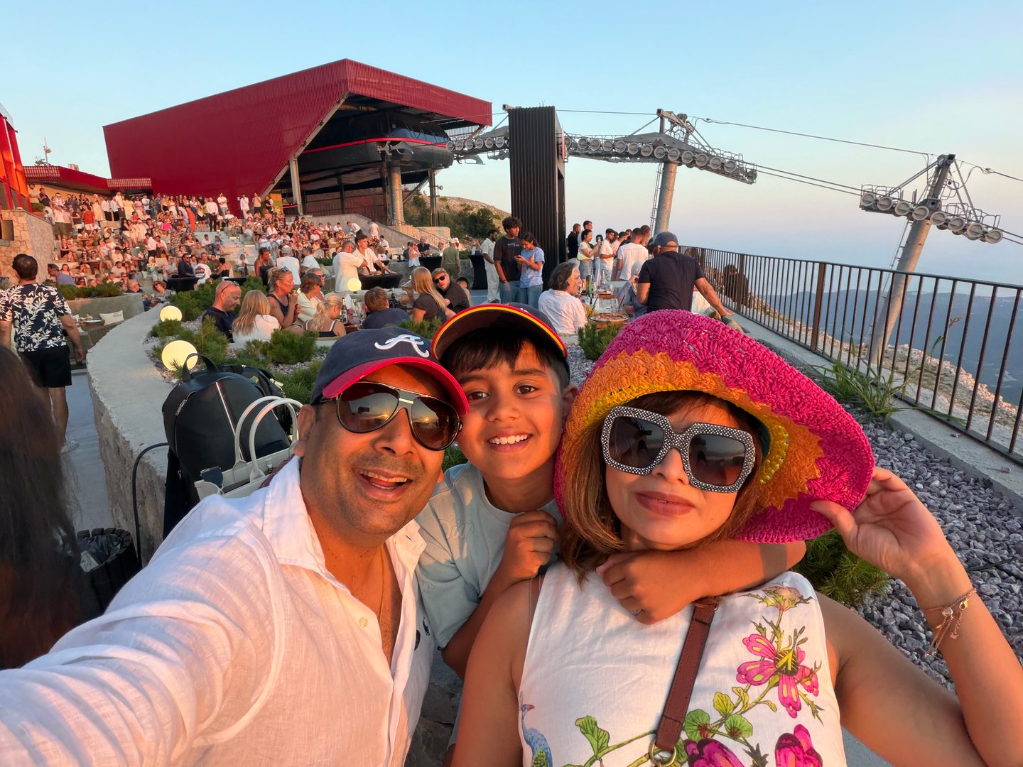 A close family selfie in warm evening light beside a busy terrace and cable-car station.